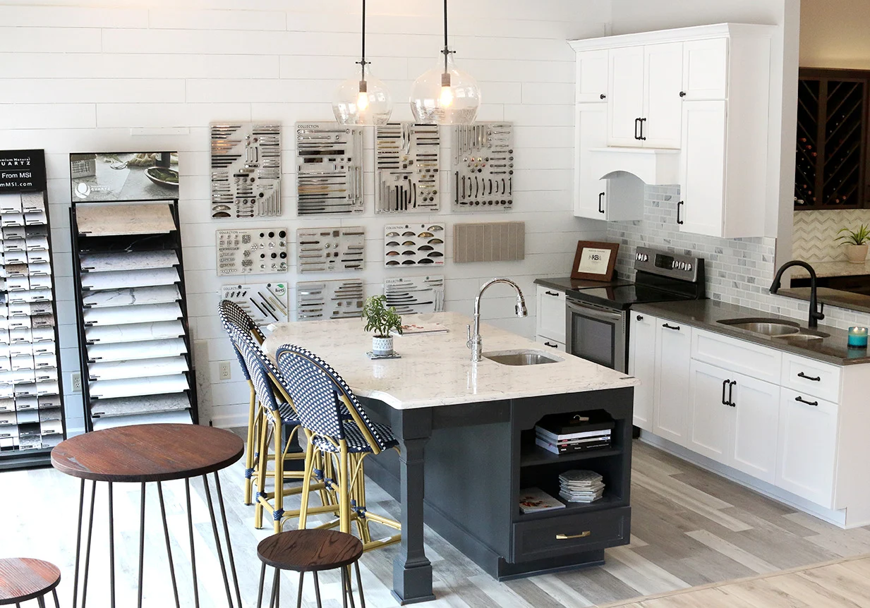 A modern kitchen showroom perfect for your kitchen remodel, featuring white cabinets, a large island with a marble countertop, blue and gold barstools, pendant lights, and display walls showcasing various cabinet handles and samples. A modern kitchen showroom perfect for your kitchen remodel, featuring white cabinets, a large island with a marble countertop, blue and gold barstools, pendant lights, and display walls showcasing various cabinet handles and samples.
