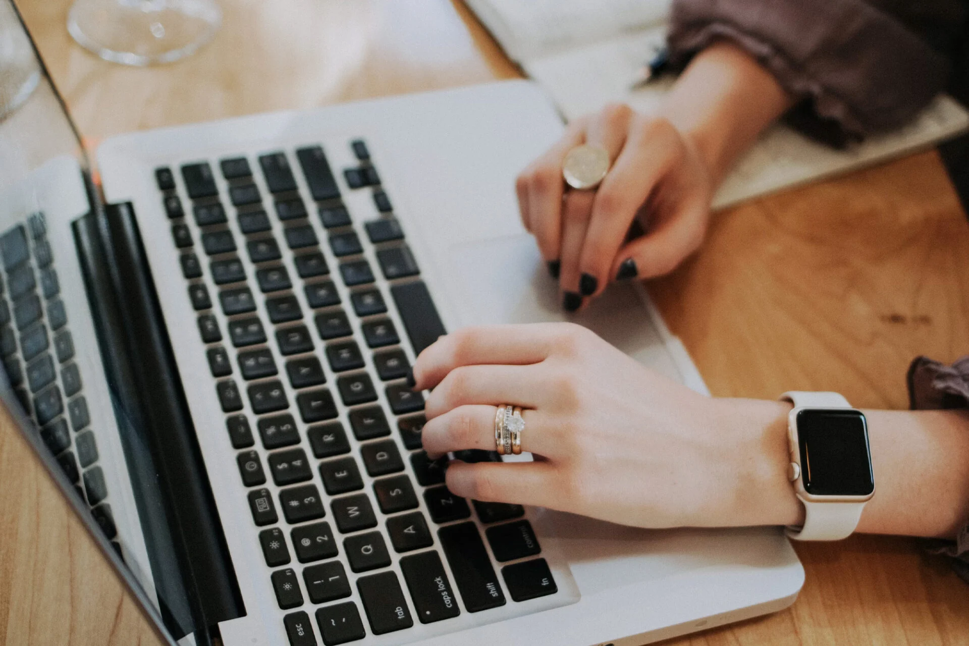A person with rings and a smartwatch types on a laptop keyboard while holding a pen, planning a kitchen remodel, with a notebook open nearby on a wooden desk. A person with rings and a smartwatch types on a laptop keyboard while holding a pen, planning a kitchen remodel, with a notebook open nearby on a wooden desk.