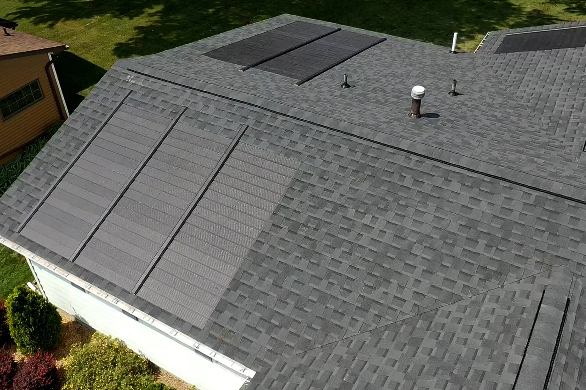 Aerial view of a gray shingled house roof featuring two sets of rectangular black solar panels and several vents, with the home’s recent kitchen remodel nestled amid surrounding trees and grass. Aerial view of a gray shingled house roof featuring two sets of rectangular black solar panels and several vents, with the home’s recent kitchen remodel nestled amid surrounding trees and grass.