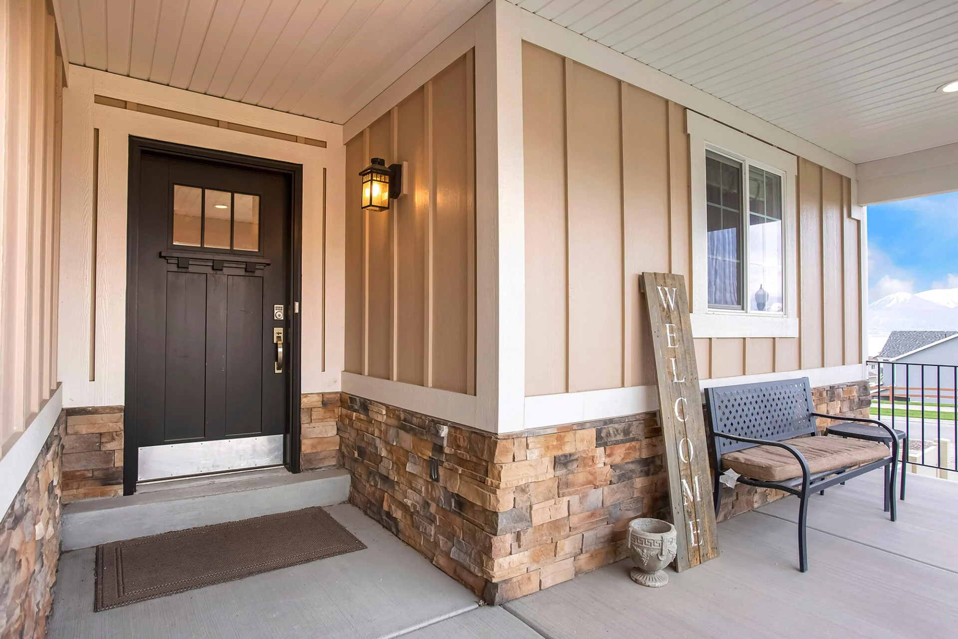 Covered front porch with stone and tan siding, a black door with a welcome mat, wall lantern, bench, and a rustic wooden WELCOME sign near the window—perfect for greeting guests after your kitchen remodel. Covered front porch with stone and tan siding, a black door with a welcome mat, wall lantern, bench, and a rustic wooden WELCOME sign near the window—perfect for greeting guests after your kitchen remodel.