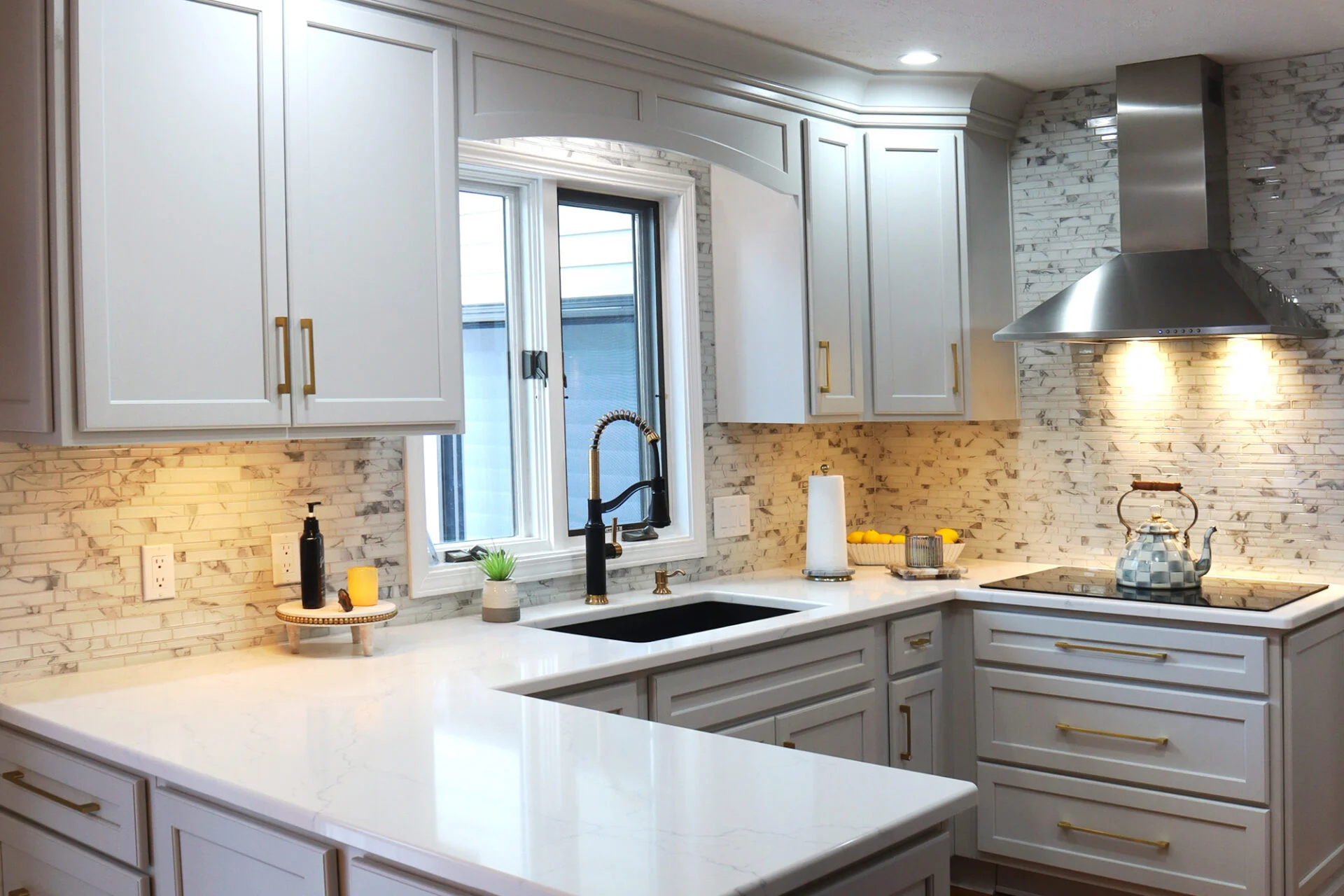 Modern kitchen remodel with white cabinets, marble backsplash, and sleek white countertops. A black sink sits under the window, near a stainless steel stove and vent hood. Small plants and kitchen items decorate the space. Modern kitchen remodel with white cabinets, marble backsplash, and sleek white countertops. A black sink sits under the window, near a stainless steel stove and vent hood. Small plants and kitchen items decorate the space.