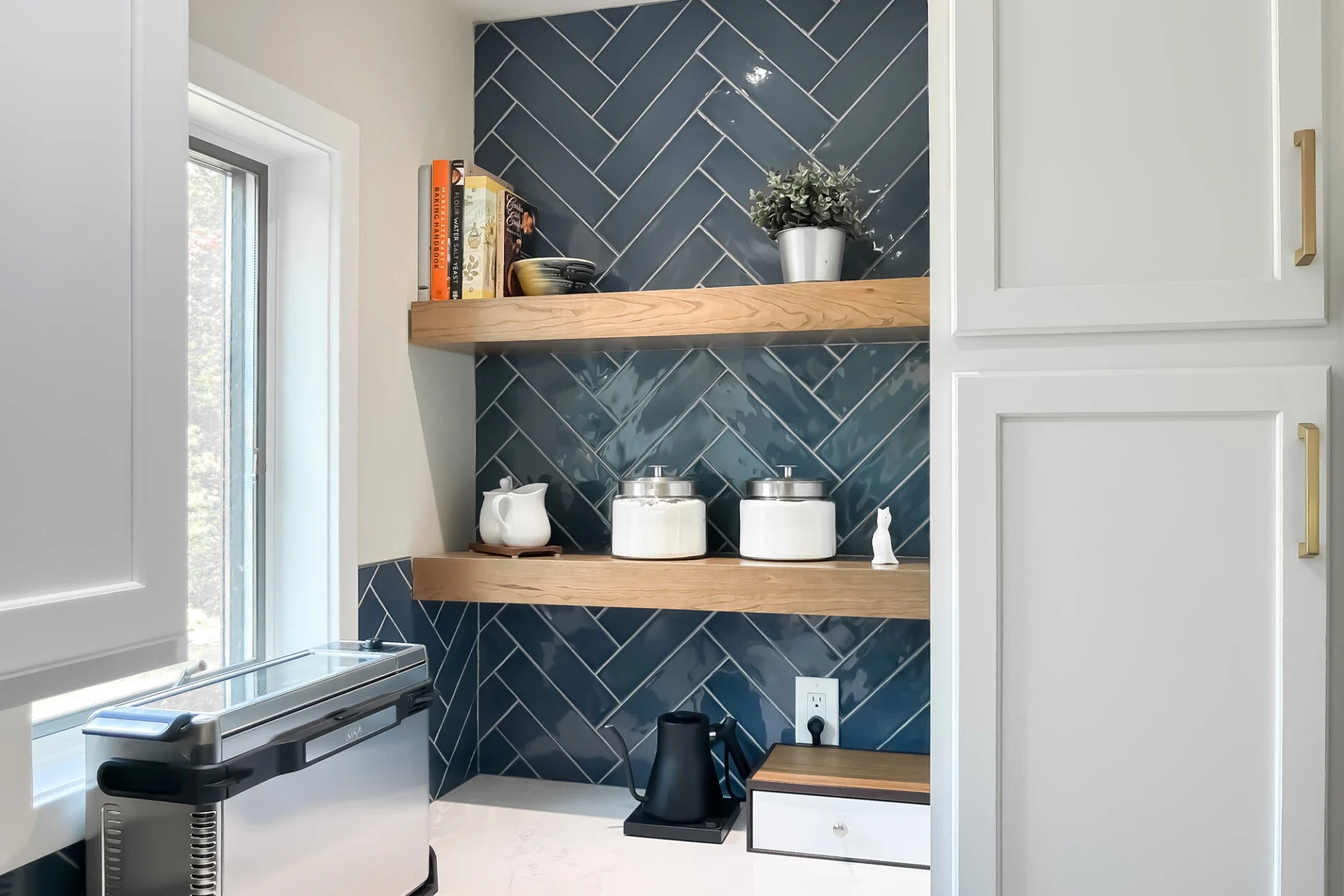 A modern kitchen remodel corner features a blue herringbone tile backsplash, two wooden shelves with cookbooks, jars, dishes, and a potted plant. A toaster, kettle, and drawer sit on the sleek white countertop below. A modern kitchen remodel corner features a blue herringbone tile backsplash, two wooden shelves with cookbooks, jars, dishes, and a potted plant. A toaster, kettle, and drawer sit on the sleek white countertop below.