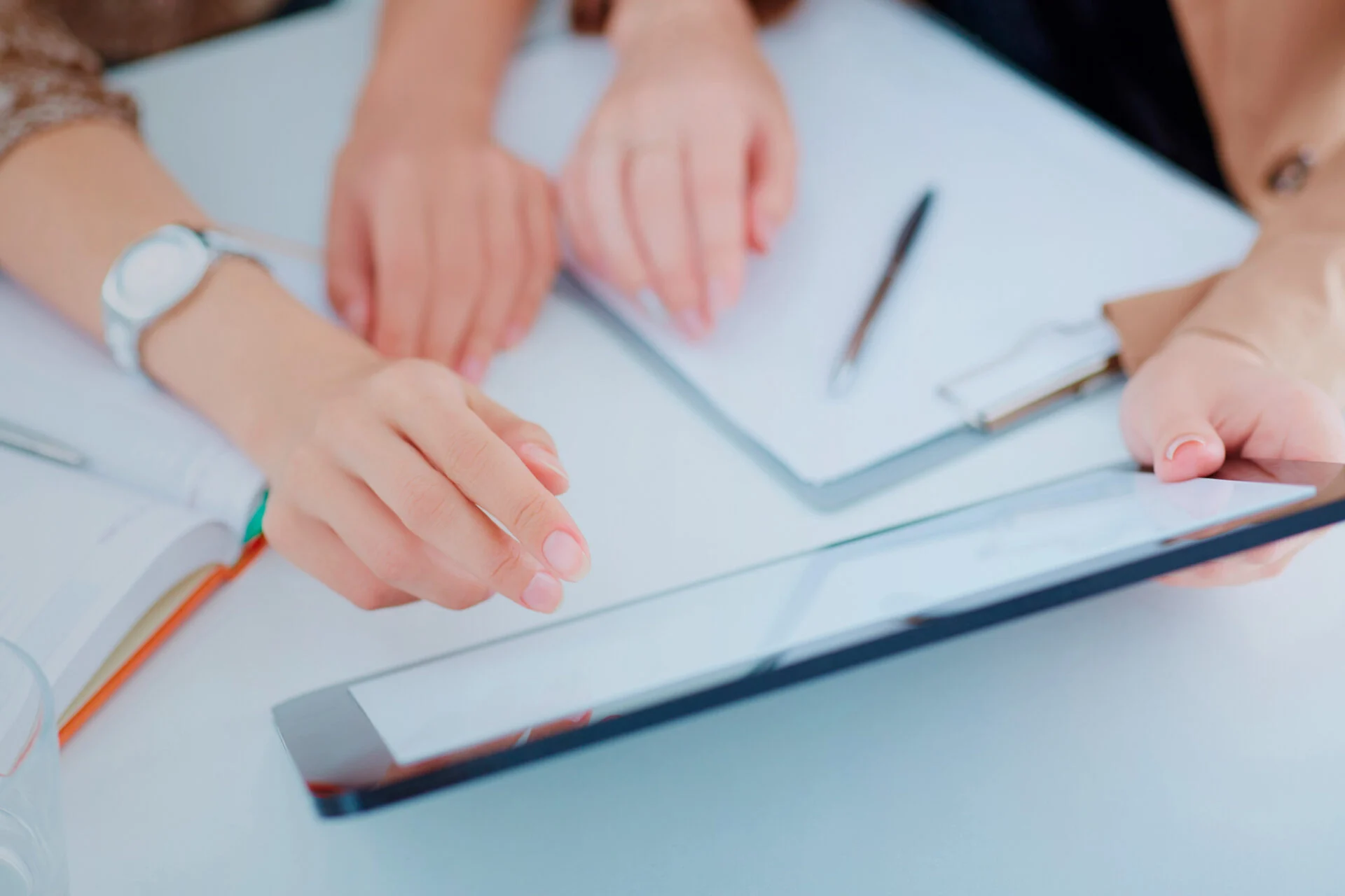 Three people sit at a table discussing a kitchen remodel with documents and a tablet. One person points at the tablet screen, while the others have hands resting near a clipboard with papers and pen on the table. Three people sit at a table discussing a kitchen remodel with documents and a tablet. One person points at the tablet screen, while the others have hands resting near a clipboard with papers and pen on the table.