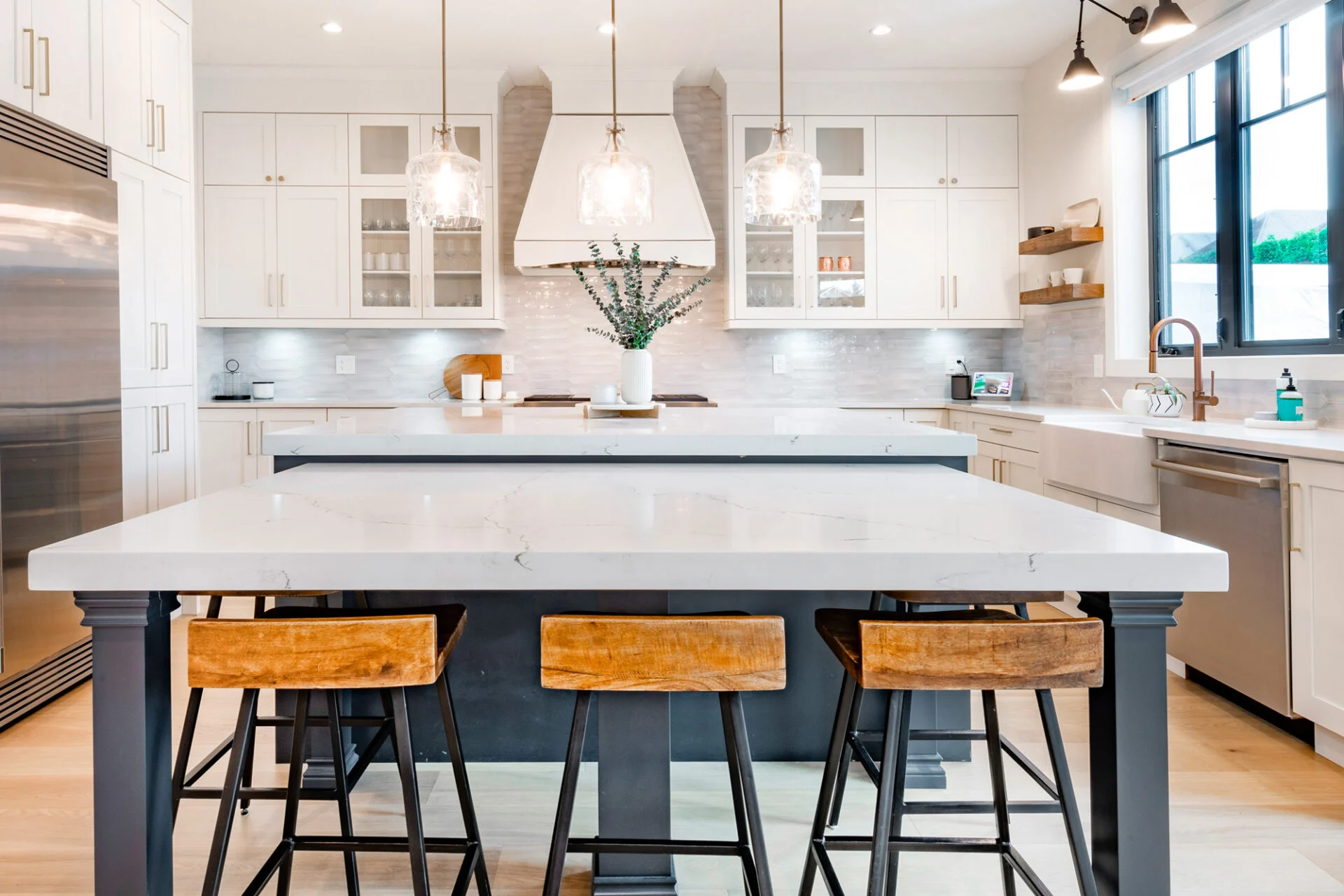 Modern kitchen remodel featuring white cabinets, a large island with a white countertop, three wooden stools, pendant lights, stainless steel appliances, and a vase with greenery on the island. Modern kitchen remodel featuring white cabinets, a large island with a white countertop, three wooden stools, pendant lights, stainless steel appliances, and a vase with greenery on the island.