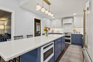 Modern kitchen remodel featuring a large white marble island, blue lower cabinets, white upper cabinets, pendant lights, a vase of flowers on the island, and sleek stainless steel appliances.