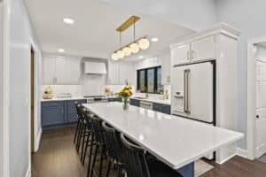 Modern kitchen remodel featuring white and blue cabinets, a large white marble island with black chairs, stainless steel appliances, a vase of flowers on the island, and a gold light fixture above.