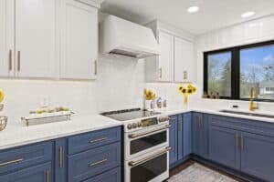 Modern kitchen remodel with white upper cabinets, blue lower cabinets, a white range hood, stainless steel oven, white tile backsplash, marble countertops, gold hardware, sunflowers in a vase, and a window with a view outside.