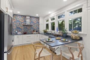 Bright kitchen remodel with white cabinets, wood floor, colorful fruit-patterned accent wall, stainless steel appliances, large windows, and a cozy dining area for two decorated with fruits and plants.