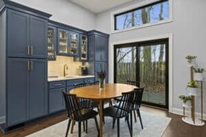 A modern kitchen remodel features blue cabinets, a wooden dining table with four black chairs, a vase of purple flowers, large windows, a sliding glass door, and a scenic view of trees outside.