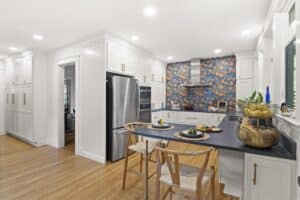 Modern kitchen remodel with white cabinets, stainless steel appliances, wood flooring, a center island with two chairs, and a blue backsplash featuring a colorful leaf pattern behind the stove. Fruit and plants decorate the countertops.