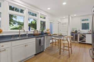 Bright, modern kitchen remodel with white cabinets, stainless steel appliances, wood floors, black countertops, large windows with outdoor views, a central island with two wooden chairs, and a built-in wine cooler in the corner.