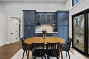 Modern kitchen remodel with blue cabinets, glass-front doors, a round wooden dining table with six black chairs, a vase of purple flowers in the center, large windows, and a scenic view of trees outside.