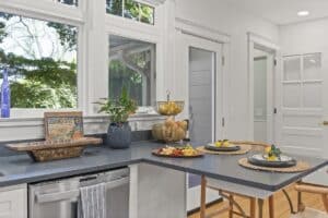 Bright kitchen with large windows, a gray countertop, fruit bowls, a plant, and breakfast plates set on a dining bar—this recent kitchen remodel lets sunlight stream in, highlighting the fresh and inviting atmosphere.