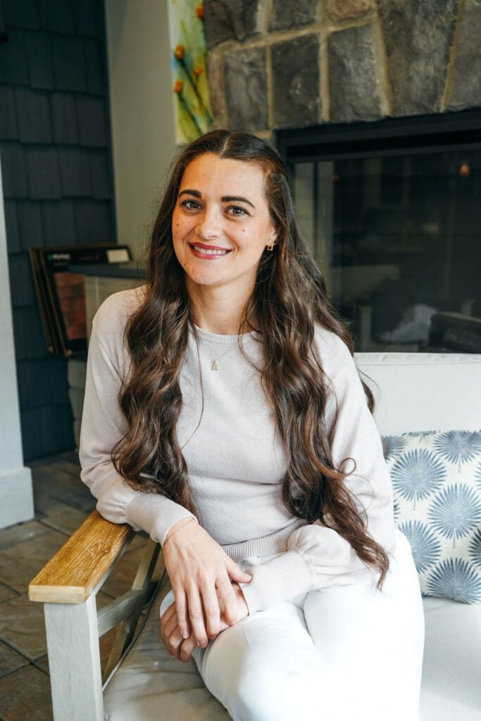 A woman with long brown hair, wearing a light sweater and white pants, sits on a chair indoors, smiling at the camera. A pillow rests beside her, with a stone fireplace and hints of a recent kitchen remodel in the background.