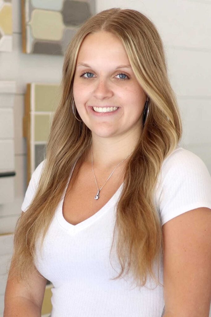 A young woman with long blonde hair, wearing a white short-sleeve top, hoop earrings, and a necklace, smiles at the camera. The background features light-colored, tiled walls.