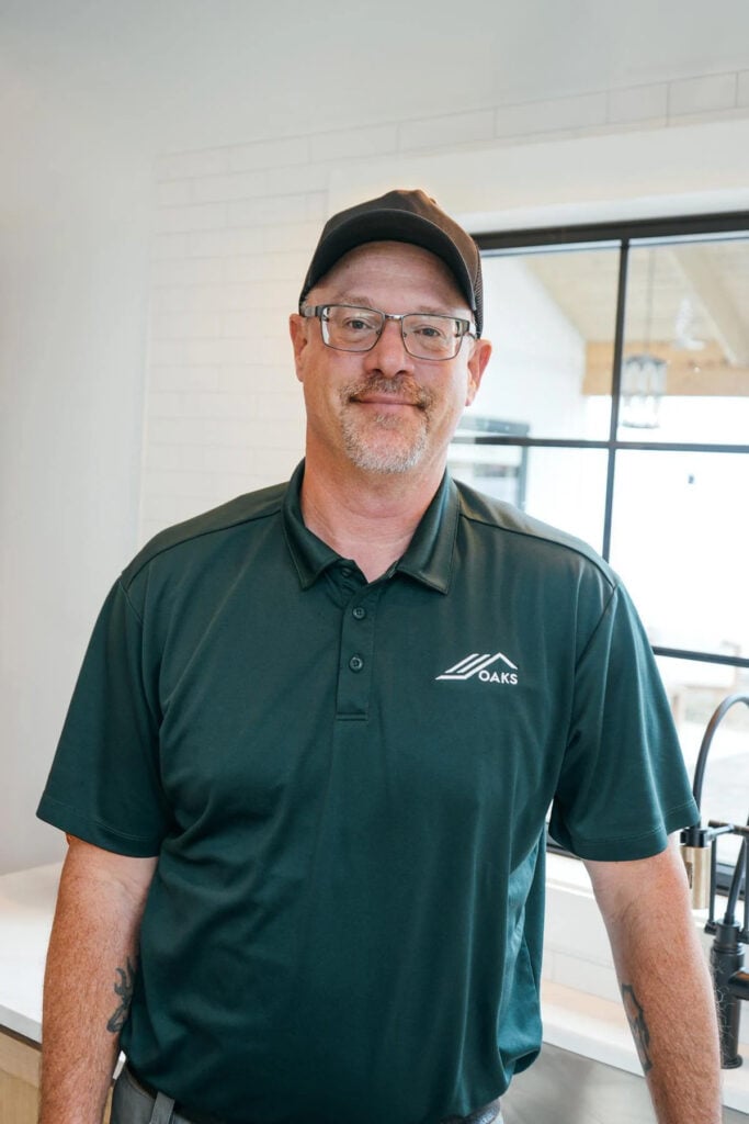 A man wearing glasses, a black cap, and a dark green polo shirt with an Oaks logo stands indoors, smiling, with a kitchen and window in the background.