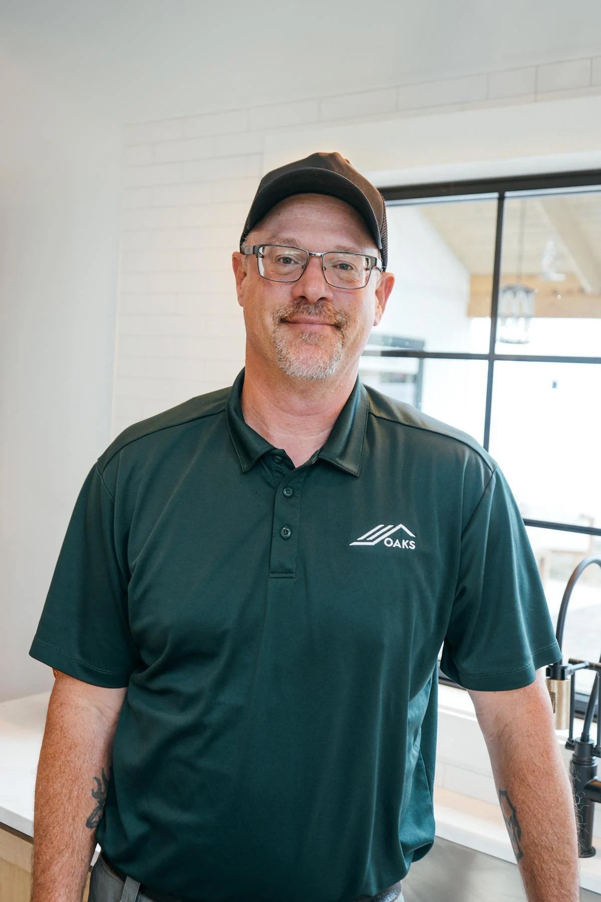 A man wearing glasses, a black cap, and a dark green polo shirt with an Oaks logo stands indoors, smiling, with a kitchen and window in the background.