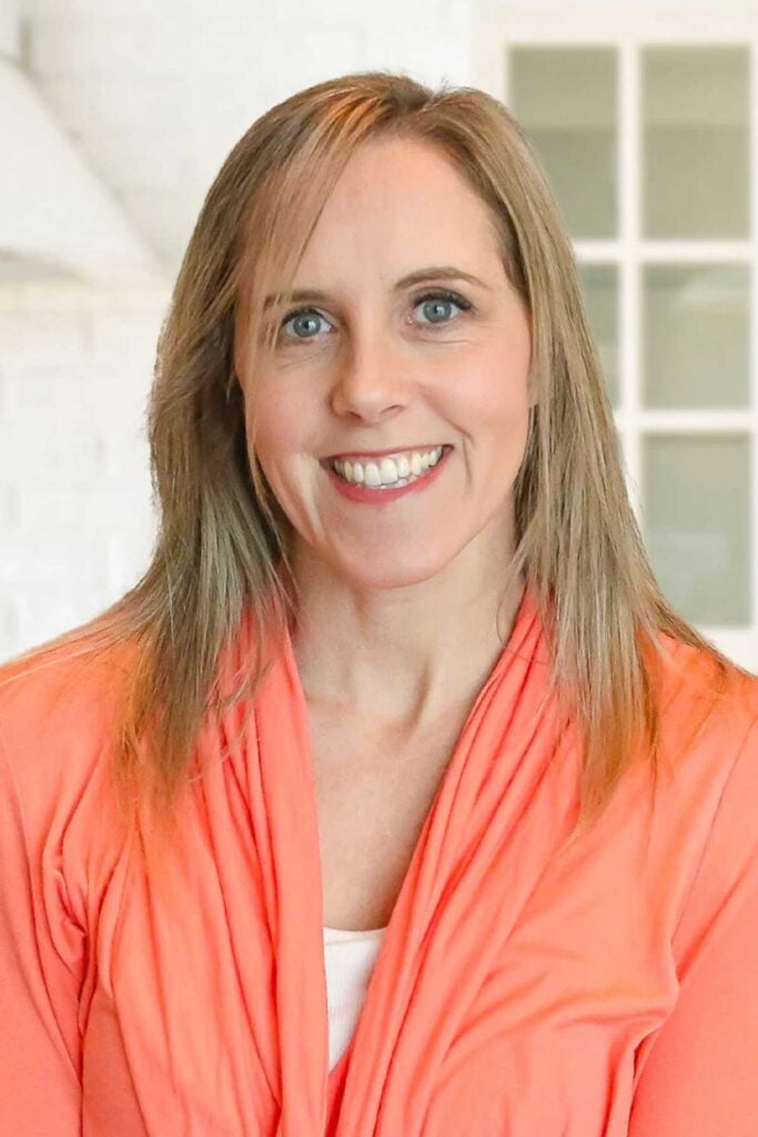 A woman with straight, light brown hair wearing a bright coral cardigan and white top smiles in a brightly lit room with white walls and a window in the background.
