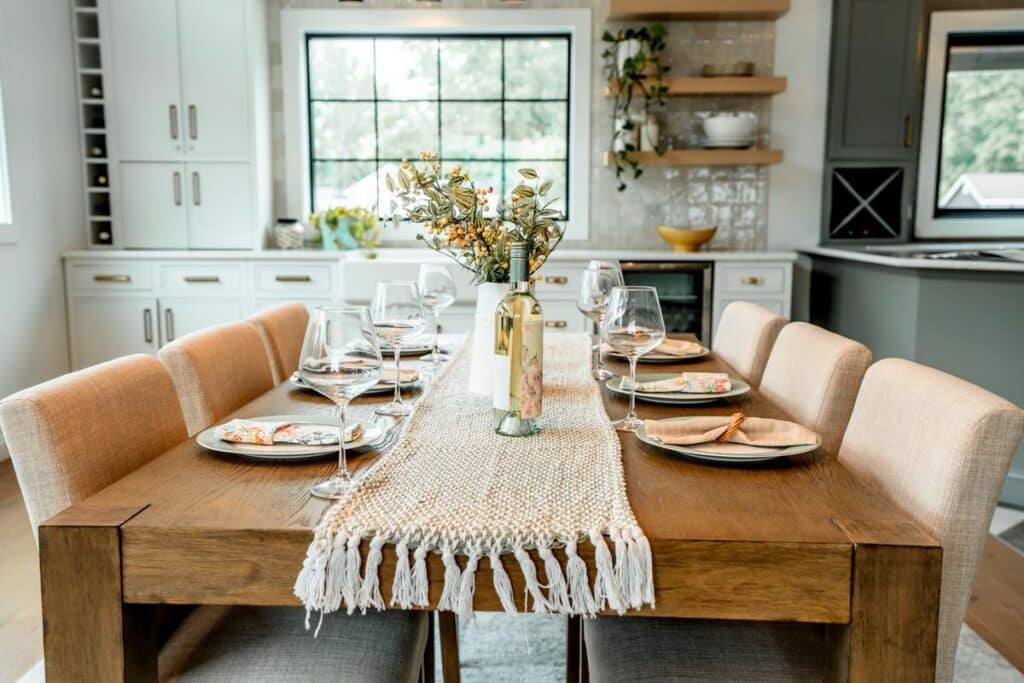 A wooden dining table set for six with wine glasses, plates, and a floral centerpiece creates a welcoming scene in a bright, modern kitchen—just like the elegant designs showcased in the Webster Showroom.