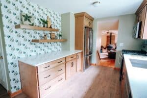 Bright kitchen remodel with light wood cabinets, white countertops, leafy patterned accent wall, and floating shelves with decor; stainless steel fridge and a view into the cozy living room in the background.