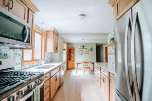 Bright kitchen remodel with wood cabinets, stainless steel appliances, a gas stove, and a sink under a window. In the background, a dining area with a wooden table and chairs sits near a large window.