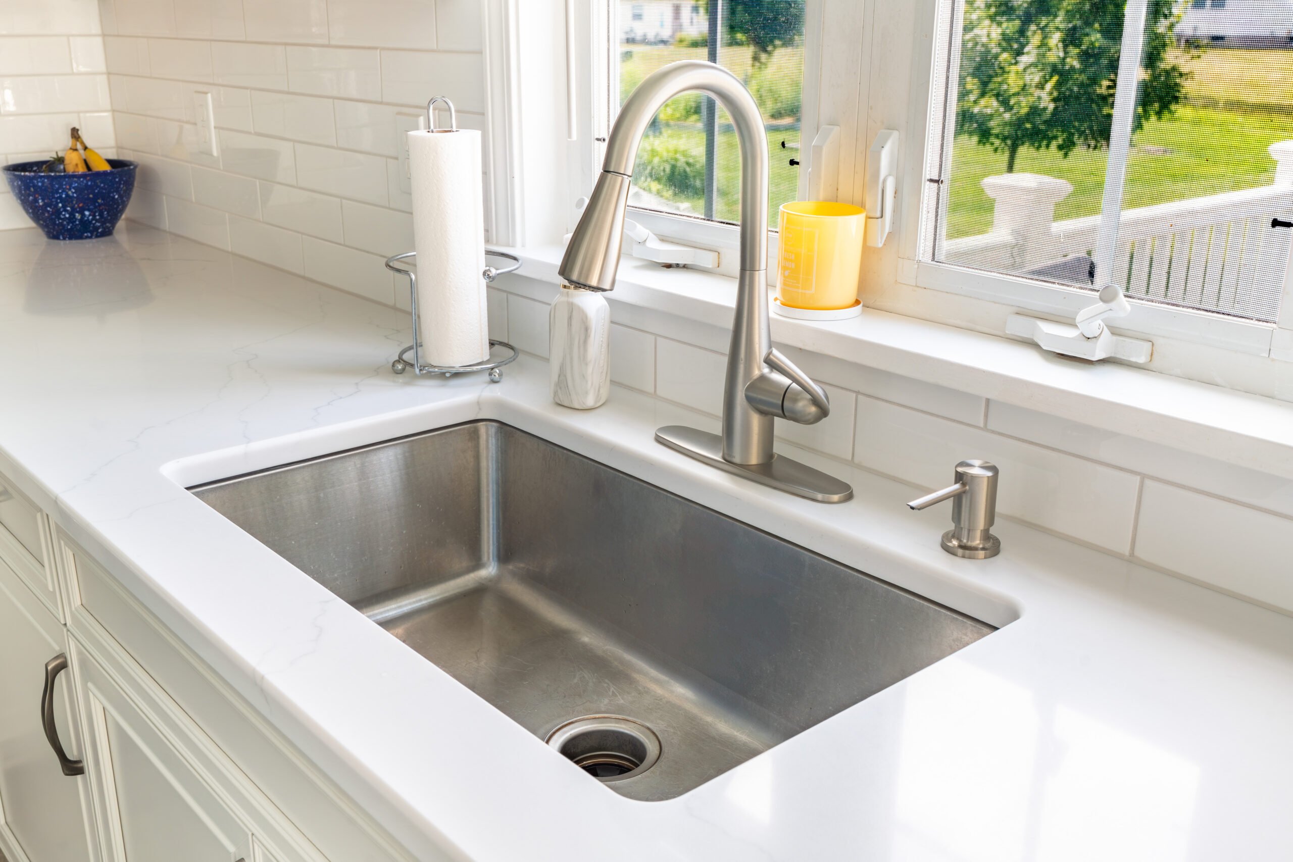 A Clean Stainless Steel Kitchen Sink With A Modern Faucet, Reverse Osmosis System, Soap Dispenser, And Paper Towel Holder On A White Countertop A Yellow Cup And Sponge Rest On The Windowsill Above, Sunlight Streaming In A clean stainless steel kitchen sink with a modern faucet, reverse osmosis system, soap dispenser, and paper towel holder on a white countertop. A yellow cup and sponge rest on the windowsill above, sunlight streaming in.