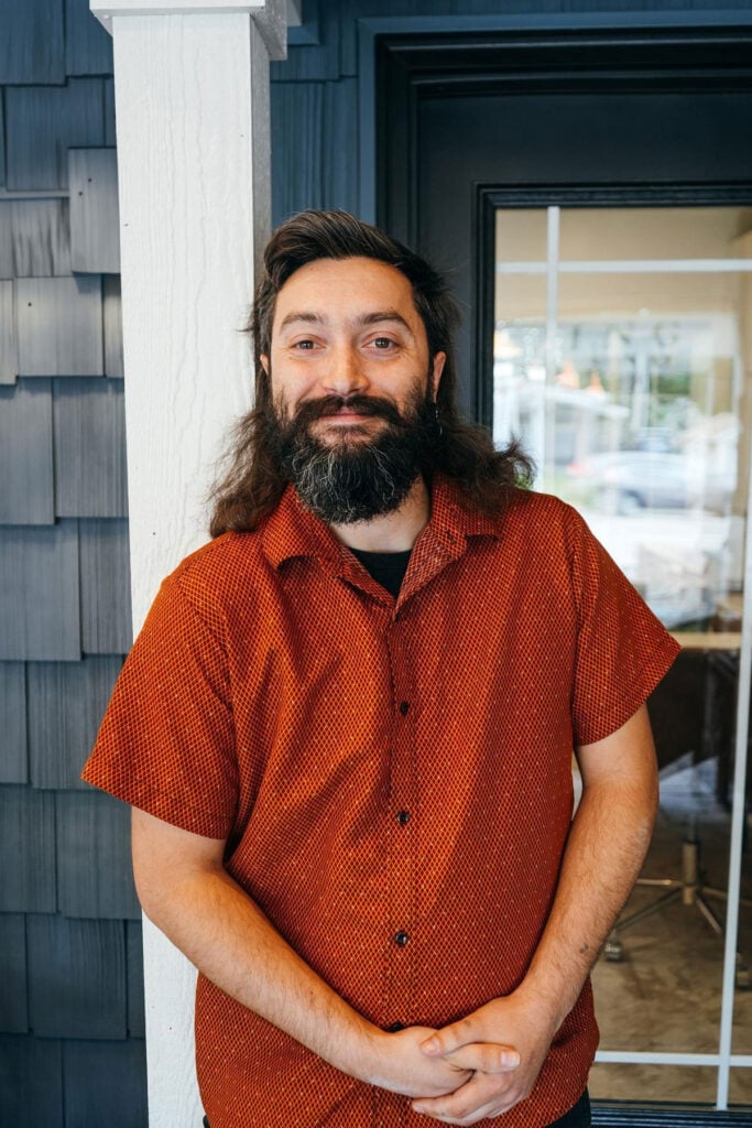 A man with long hair and a beard smiles while standing outside a building with dark siding and a glass door. He is wearing a short-sleeved, red patterned shirt and has his hands folded in front of him.