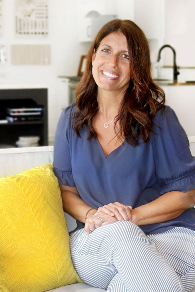 A woman with long brown hair, wearing a blue blouse and striped pants, sits on a light-colored couch with a bright yellow pillow. She smiles in a modern, well-lit living room that beautifully complements her recent kitchen remodel.