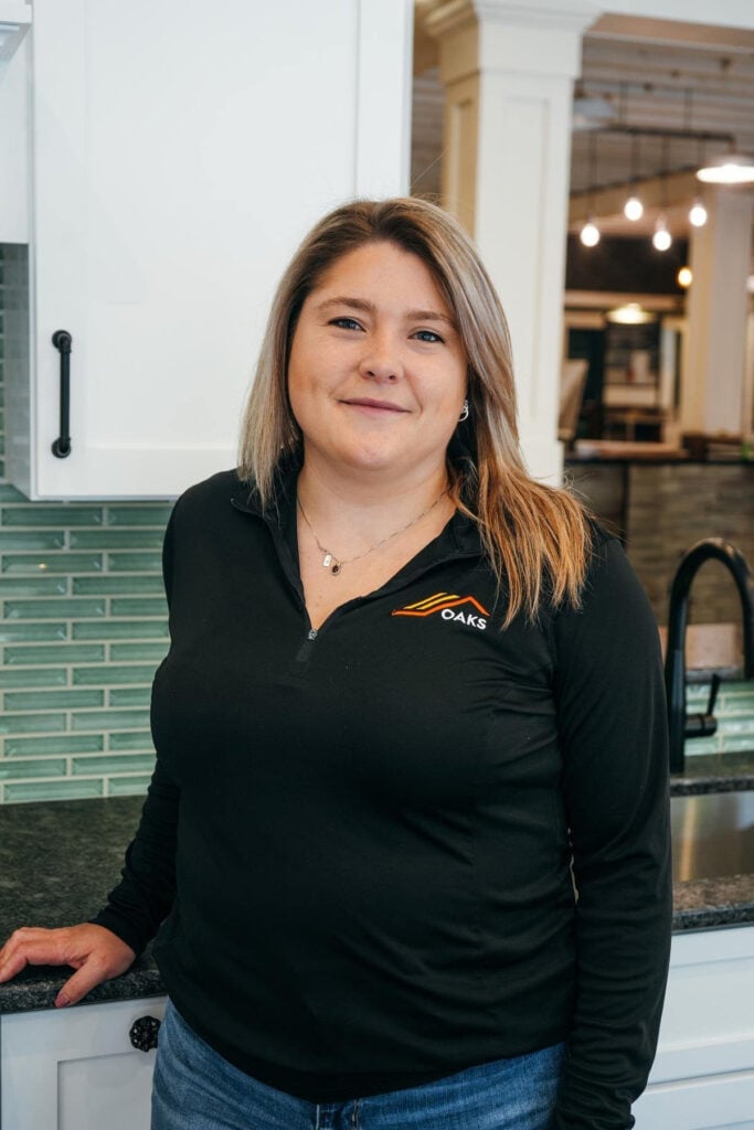 A woman with light brown hair wearing a black long-sleeve shirt with the OAKS logo stands in a modern kitchen, smiling at the camera. Green tile backsplash and white cabinets are visible in the background.