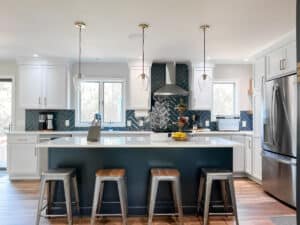 Modern kitchen with white cabinets, stainless steel appliances, blue tile backsplash, and a large island with four metal stools. Three pendant lights hang above the island, and a bowl of fruit sits on the countertop.