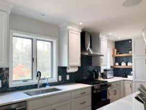 Modern kitchen with white cabinets, stainless steel appliances, a herringbone-patterned dark tile backsplash, and a large window over the sink letting in natural light. Countertops are white with minimal items placed neatly.