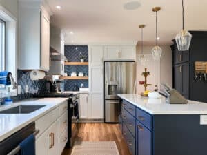 Modern kitchen with white and navy cabinets, stainless steel appliances, a patterned tile backsplash, pendant lights over a white island with a fruit stand, and wooden floor. Natural light comes in through a window above the sink.