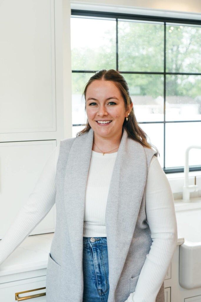 A woman with long brown hair, wearing a white long-sleeve top and a light gray vest, stands smiling in a bright kitchen with large windows in the background.
