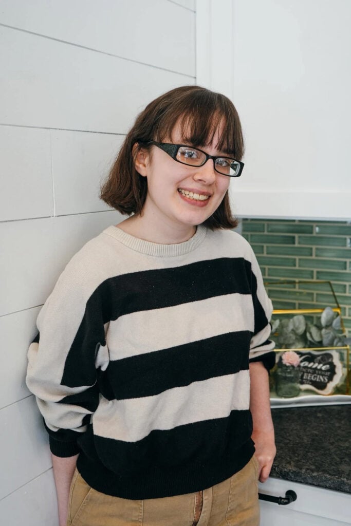 A person with short brown hair and glasses, wearing a black and white striped sweater and tan pants, smiles while leaning against a white wall in a kitchen with green tile backsplash.