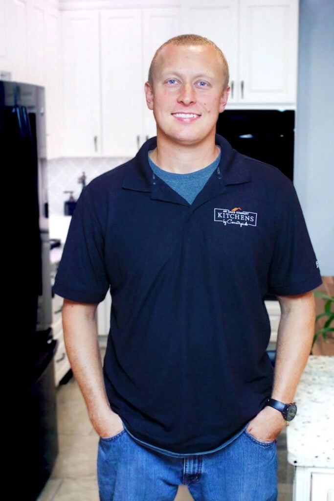 A smiling man with short blond hair stands in a kitchen, wearing a black polo shirt with a Kitchens by Countryside logo, blue jeans, and a watch, with his hands in his pockets.