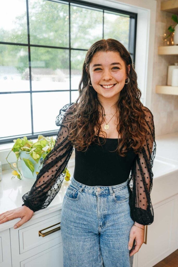 Young woman with long wavy brown hair, wearing a sheer black polka dot blouse and high-waisted jeans, smiles while standing in a bright kitchen with a large window and plants on the counter.