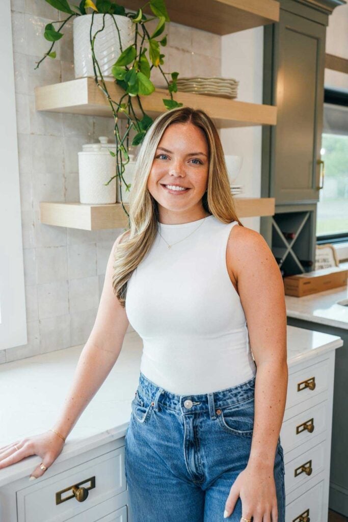 A woman with long blonde hair, wearing a white sleeveless top and blue jeans, stands in a modern kitchen with white countertops, green cabinets, and wooden shelves with plants and jars. She is smiling at the camera.