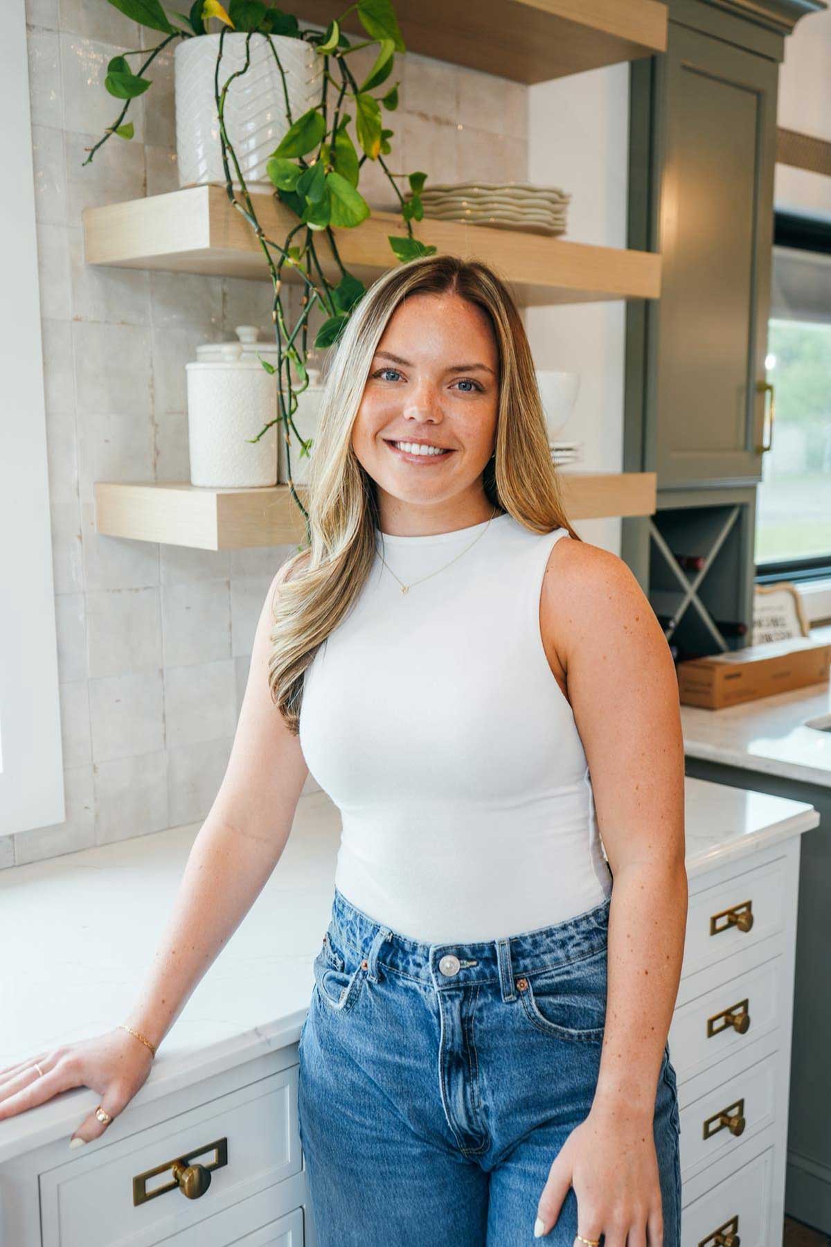 A woman with long blonde hair, wearing a white sleeveless top and blue jeans, stands in a modern kitchen with white countertops, green cabinets, and wooden shelves with plants and jars. She is smiling at the camera. A woman with long blonde hair, wearing a white sleeveless top and blue jeans, stands in a modern kitchen with white countertops, green cabinets, and wooden shelves with plants and jars. She is smiling at the camera.