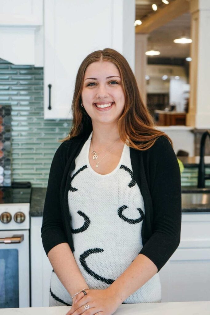 A young woman with long brown hair smiles while standing in a modern kitchen with white cabinets, a black countertop, and a green tile backsplash. She is wearing a white top with black patterns and a black cardigan.