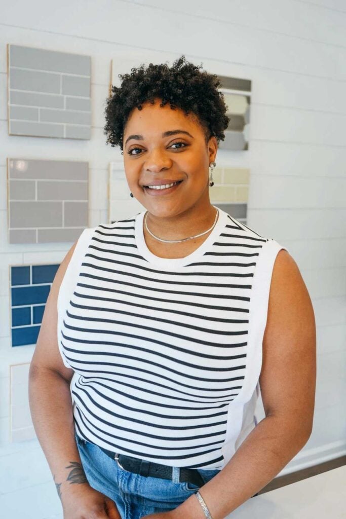 A person with short curly hair, wearing a sleeveless black and white striped top and blue jeans, smiles while standing in front of a wall displaying various tile samples in neutral colors.