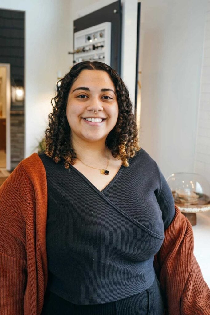 A woman with curly hair smiles at the camera. She is wearing a black top, a rust-colored cardigan, and a necklace, standing indoors in a modern, well-lit room.