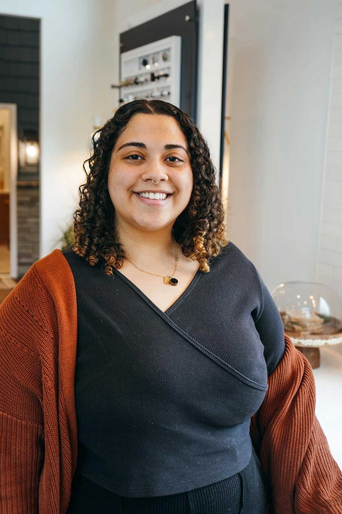 A woman with curly hair smiles at the camera. She is wearing a black top, a rust-colored cardigan, and a necklace, standing indoors in a modern, well-lit room. A woman with curly hair smiles at the camera. She is wearing a black top, a rust-colored cardigan, and a necklace, standing indoors in a modern, well-lit room.