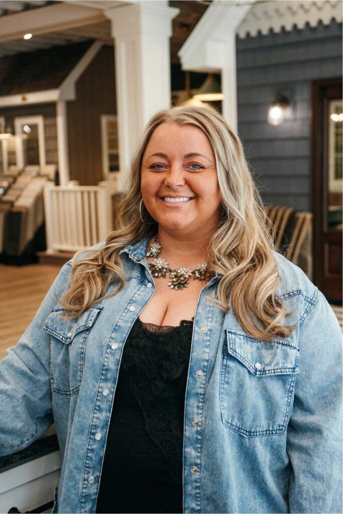 A woman with long, wavy blonde hair is smiling. She wears a denim shirt over a black lace top and a statement necklace. She stands indoors in a room with wood and neutral décor in the background.