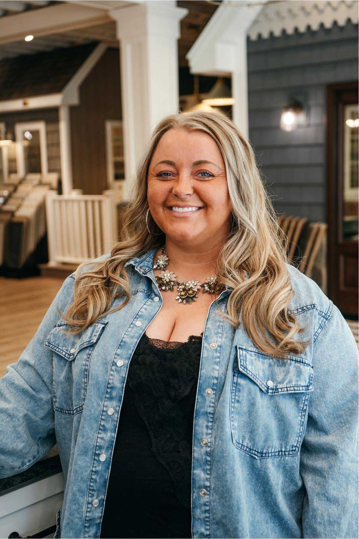 A woman with long, wavy blonde hair is smiling. She wears a denim shirt over a black lace top and a statement necklace. She stands indoors in a room with wood and neutral décor in the background. A woman with long, wavy blonde hair is smiling. She wears a denim shirt over a black lace top and a statement necklace. She stands indoors in a room with wood and neutral décor in the background.