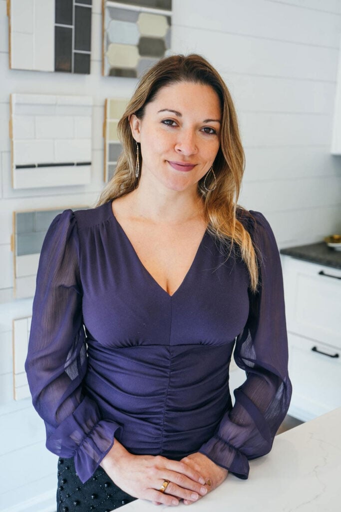 A woman with long light brown hair wearing a purple blouse stands in a modern, bright room with geometric tile samples on the wall behind her. She is smiling gently and resting her hands on a white counter.