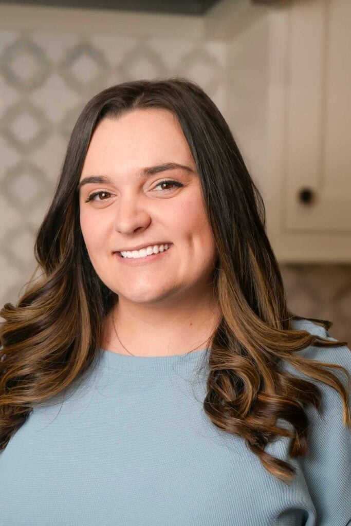 A woman with long, wavy brown hair, wearing a light blue top, smiles at the camera. The background features a light-colored kitchen with patterned tiles and cabinets.