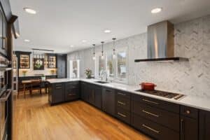 Modern kitchen remodel with dark cabinets, white countertops, stainless steel appliances, a range hood, and wood flooring. Pendant lights hang above the sink, and a dining area is visible in the background by large windows.