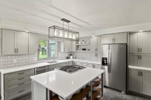 Modern kitchen remodel with gray cabinets, white countertops, stainless steel appliances, hexagonal tile backsplash, island cooktop, open shelving, and rectangular chandelier. Large window provides natural light and a view of the green yard.