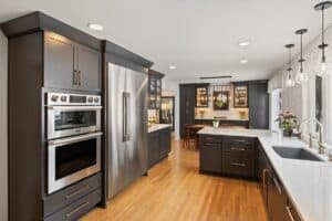 Modern kitchen remodel with dark cabinets, stainless steel appliances, a large refrigerator, built-in oven, white countertops, light wood flooring, hanging pendant lights, and a dining area in the background with a glass cabinet.