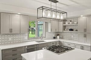 Modern kitchen remodel with light gray cabinets, geometric tile backsplash, white countertops, a gas cooktop on an island, stainless steel appliances, open shelves with decor, and a large window letting in natural light.